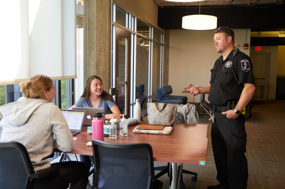 A campus police officer standing in a brightly lit, modern office space, smiling and interacting with two young women seated at a conference table with laptops and personal items.